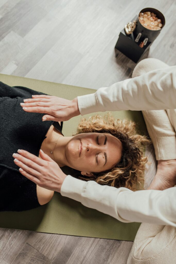 Nghi Thức Tối: 10 Phút Để Trở Về Với Chính Mình A woman receiving Reiki therapy with hands hovering over her, eyes closed, in a serene indoor setting.